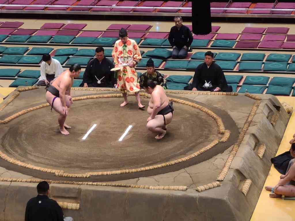 The first bout is about to erupt! You can tell these are lower-division wrestlers because the referee (gyoji) wears a simple robe, his legs are exposed, and he is barefoot. As referees rise in rank, their robes become dazzlingly complex, and their footwear upgrades from bare feet, to tabi socks, and finally to formal sandals. The absolute highest-ranking chief referee even carries a dagger, symbolizing his historical vow to commit seppuku if he ever makes a bad call!